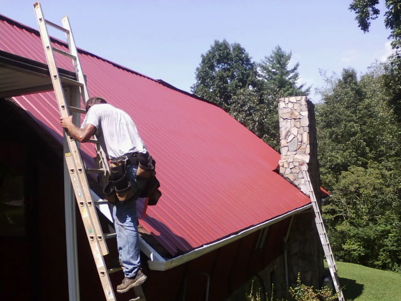 Roofer on a ladder beside a red metal roof and stone chimney — Soffit & Fascia Repair by Roofing Brothers in Hubert
